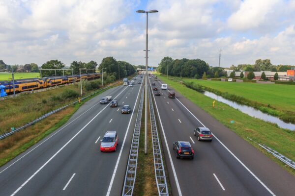 Drukke snelweg met naastgelegen spoorlijn waar een NS-trein rijdt door een groen, landelijk gebied.